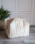 A quilted travel toiletry bag with a cream and pale pink floral pattern sits on a gray wood floor in front of a white paneled wall, next to a potted green plant.