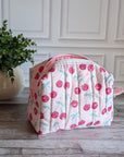 A quilted cosmetic bag with a pink and white cherry pattern, placed on a gray wood floor next to a potted green plant against a white paneled wall.