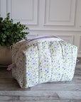 A quilted cosmetic bag with a white background and a purple and green floral pattern, placed on a gray wood floor next to a potted green plant.