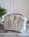 A quilted cosmetic bag with a white, purple and green floral pattern and purple gingham lining, placed on a gray wood floor next to a potted green plant.