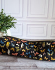 A long cylindrical black zipper pouch with a colorful butterfly print, resting on a gray wood floor next to a potted green plant against a white paneled wall.
