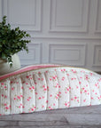 A quilted fabric makeup bag with a pink floral print and a pink zipper, resting on a gray wooden floor next to a potted green plant against a white paneled wall.