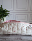 A quilted cream-colored pencil case with a pink cherry pattern and a pink gingham lining, resting on a gray wooden floor next to a potted green plant.