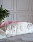 A quilted fabric zippered pouch with a cream background, small pink cherry pattern, and a pink gingham lining, resting on a gray wood floor next to a potted green plant.