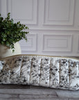 A quilted hair tool pouch with a black floral print on a white background, resting on a gray wooden floor in front of a white paneled wall.
