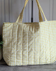 A large quilted tote bag in a pale yellow and white gingham check pattern, hanging on a black stand in front of a white paneled wall with a potted green plant on the left.