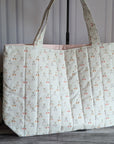 A quilted fabric tote bag with a repeating cherry print, standing on a dark wood floor against a paneled wall with a potted plant on the left.