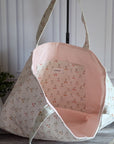 A quilted fabric tote bag with a repeating cherry print, standing on a dark wood floor against a paneled wall with a potted plant on the left.