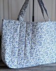 A large quilted tote bag with a blue and white floral print, standing on a wooden floor next to a potted green plant.