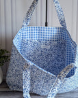 A large fabric tote bag with a blue and white floral print, standing on a gray wood floor next to a potted green plant against a paneled cream wall.