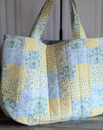 A large quilted patchwork tote bag with yellow, light blue, and white patterned fabric panels, featuring two shoulder straps, displayed on a wooden floor next to a potted plant.