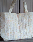 A large quilted fabric tote bag with a delicate pastel floral and bow pattern, standing on a dark wood floor next to a potted green plant.