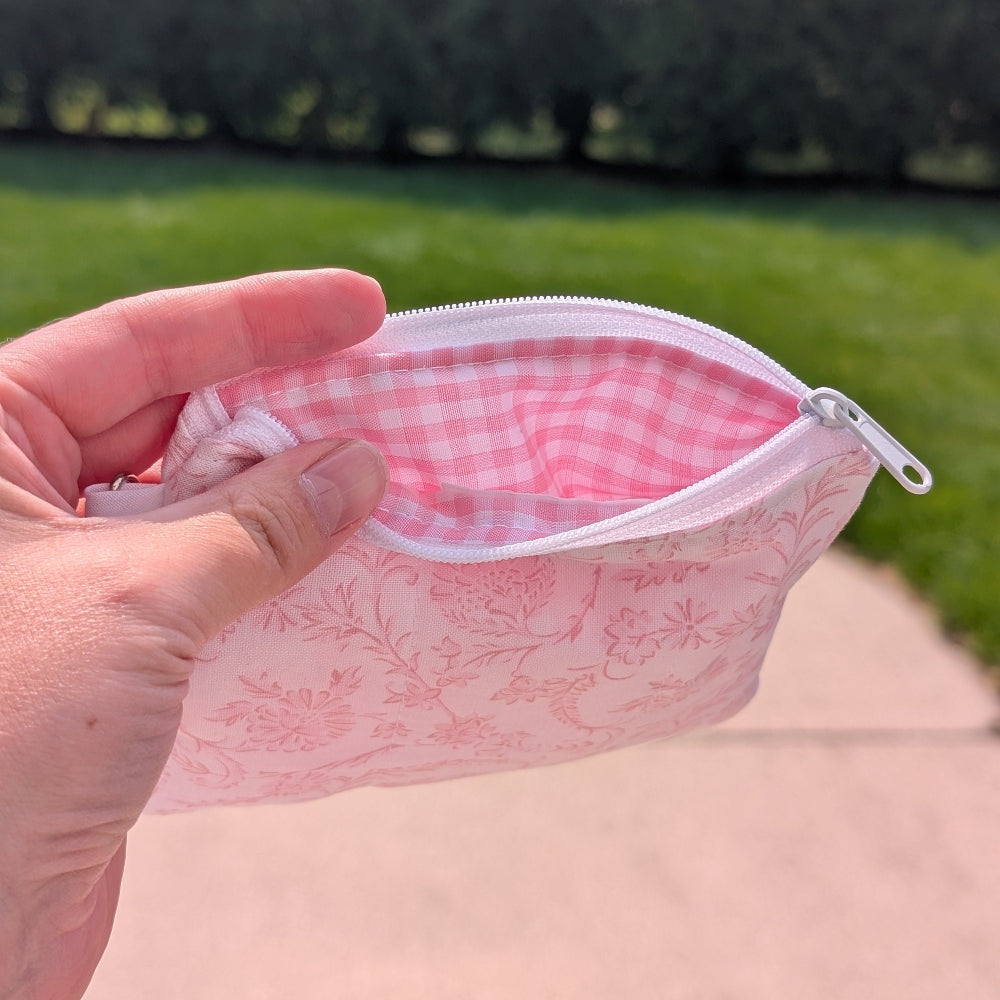 Hand holding a pink floral pouch with a checkered interior against a blurred natural background