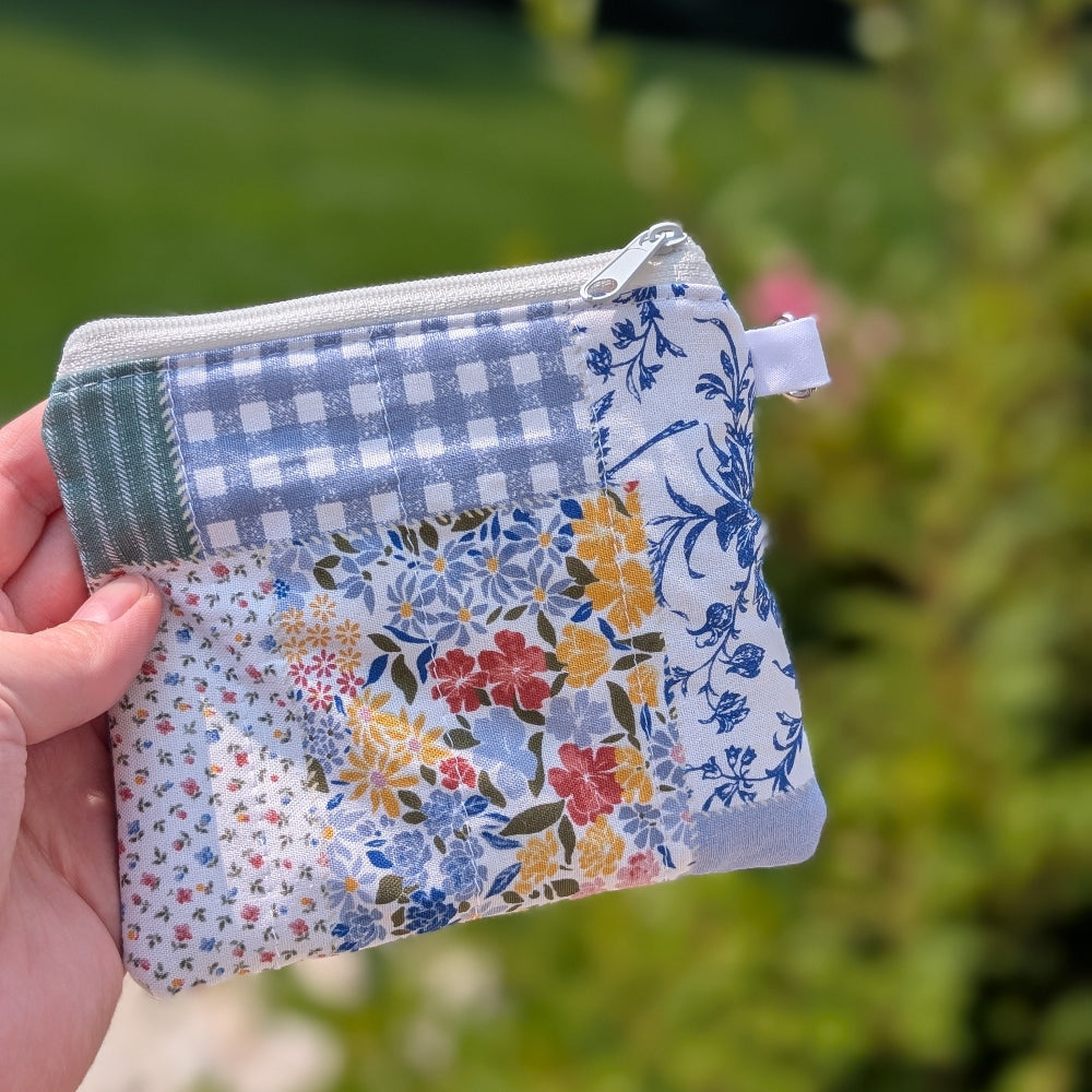 Hand holding a small fabric pouch with floral and checkered patterns against a blurred green outdoor background.
