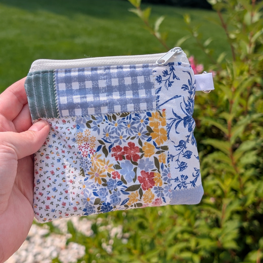 Hand holding a small fabric pouch with floral and checkered patterns against a green outdoor background.