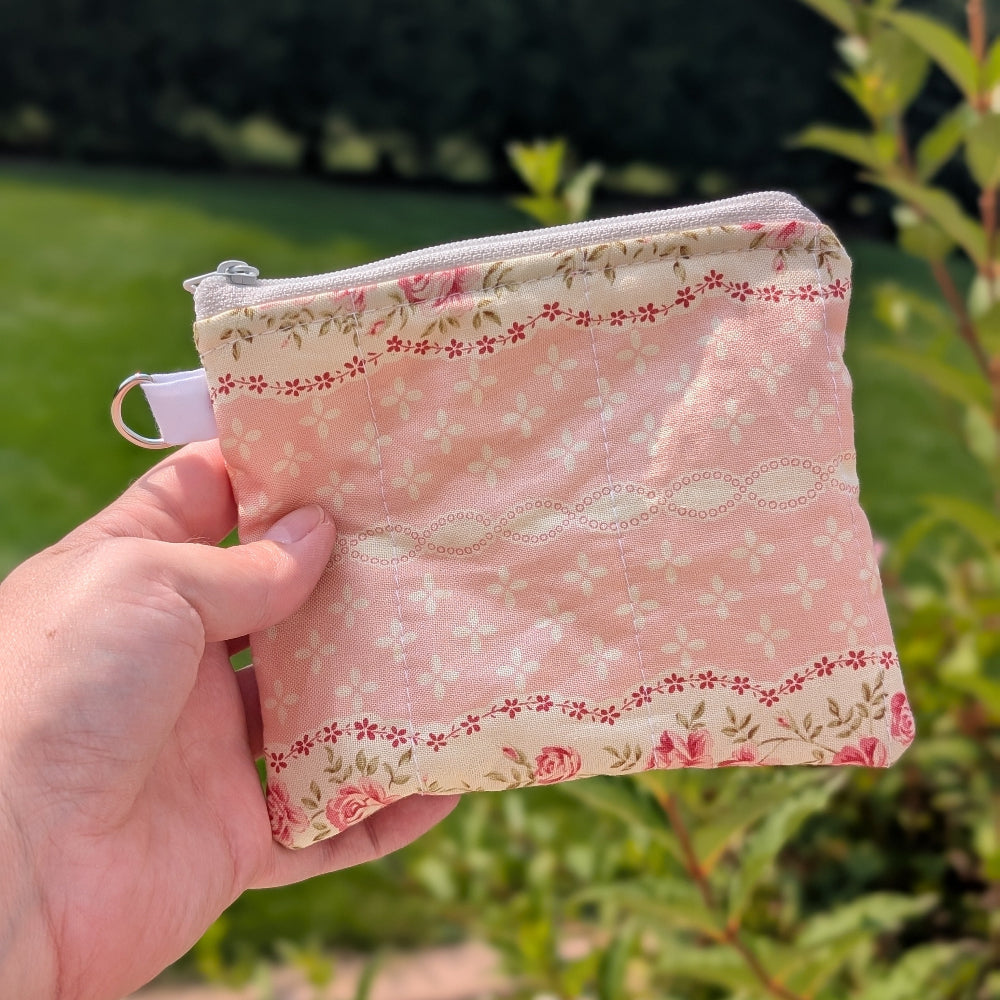 Hand holding a small pink fabric pouch with floral patterns against a green outdoor background