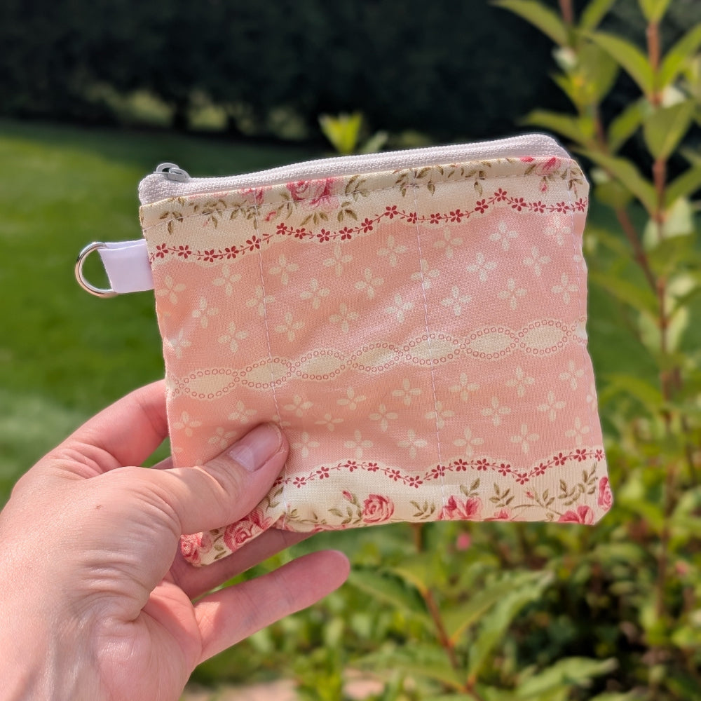 Hand holding a small pink fabric pouch with floral patterns against a green outdoor background