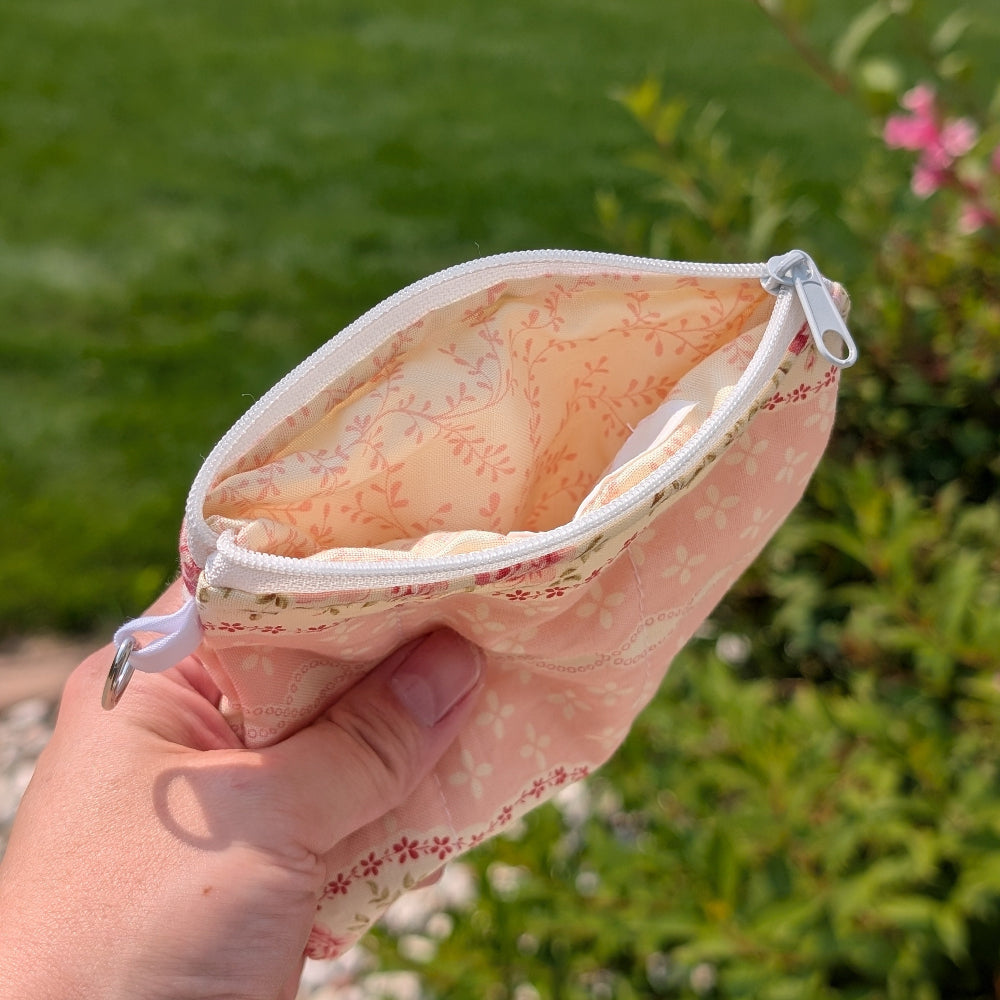 Hand holding a small pink pouch with floral pattern against a blurred green outdoor background