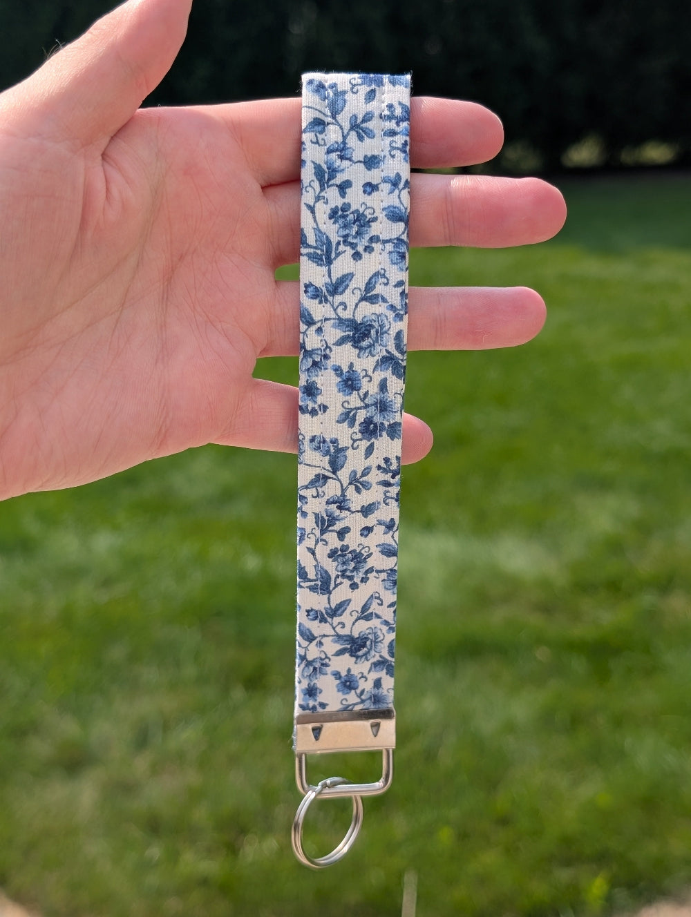 Hand holding a blue floral keychain strap against a grassy background