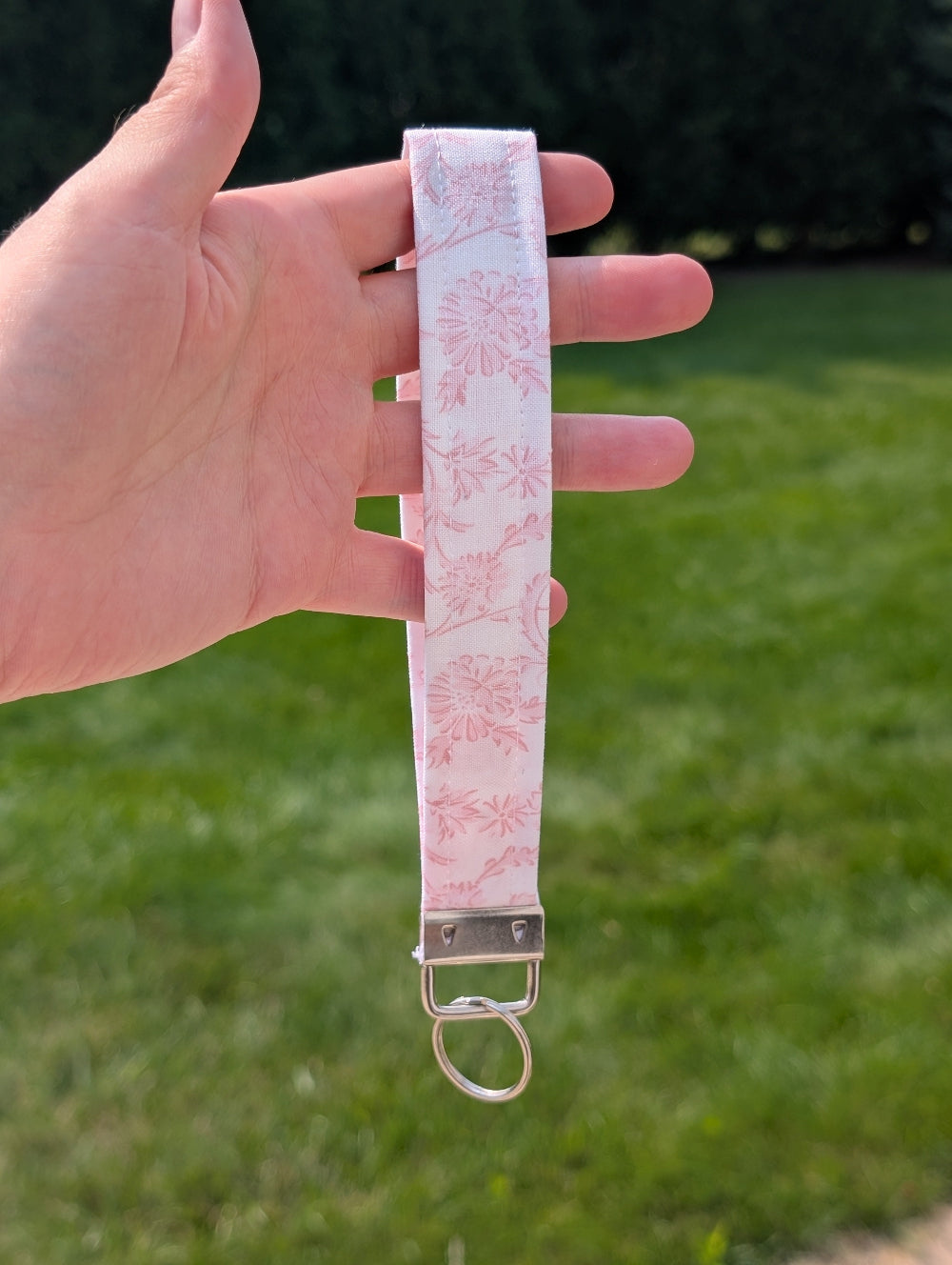 Hand holding a pink floral keychain strap with a metal clasp against a grassy background