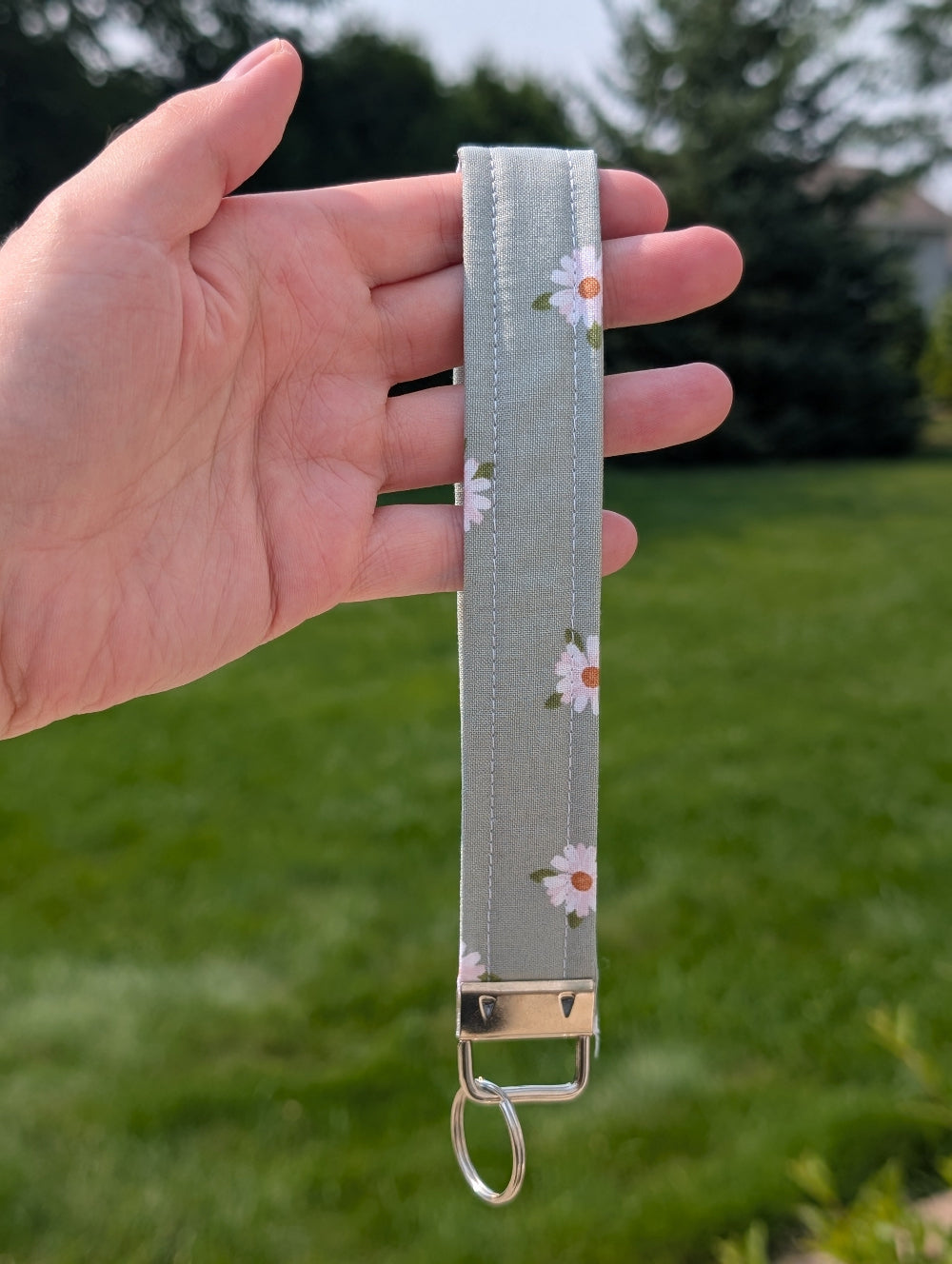 Hand holding a keychain strap with floral pattern against a green outdoor background