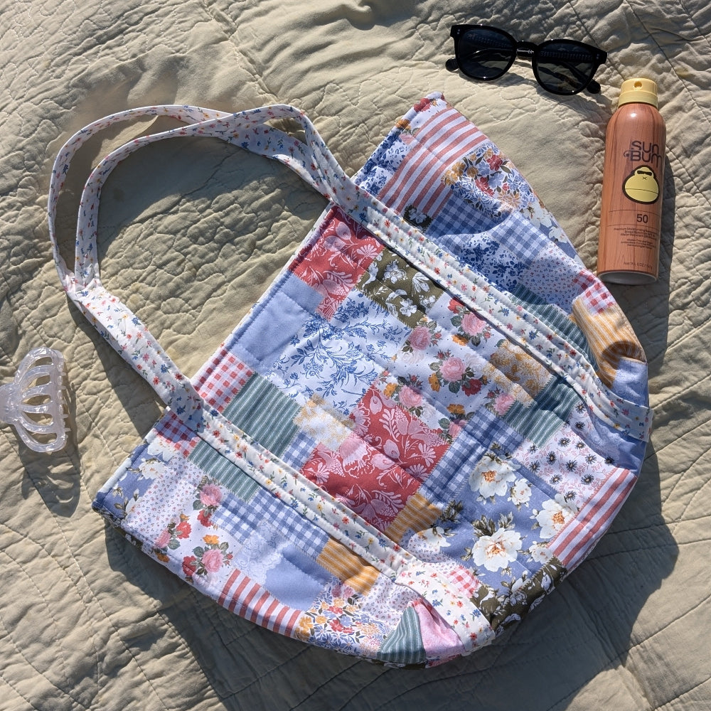 Patchwork handbag on sand with sunglasses and sunscreen