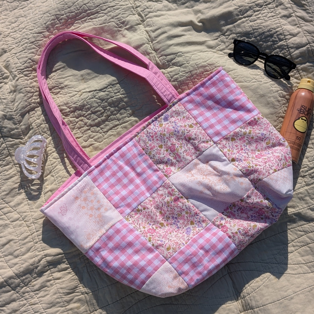 Pink patchwork handbag on a textured surface with sunglasses and a bottle.