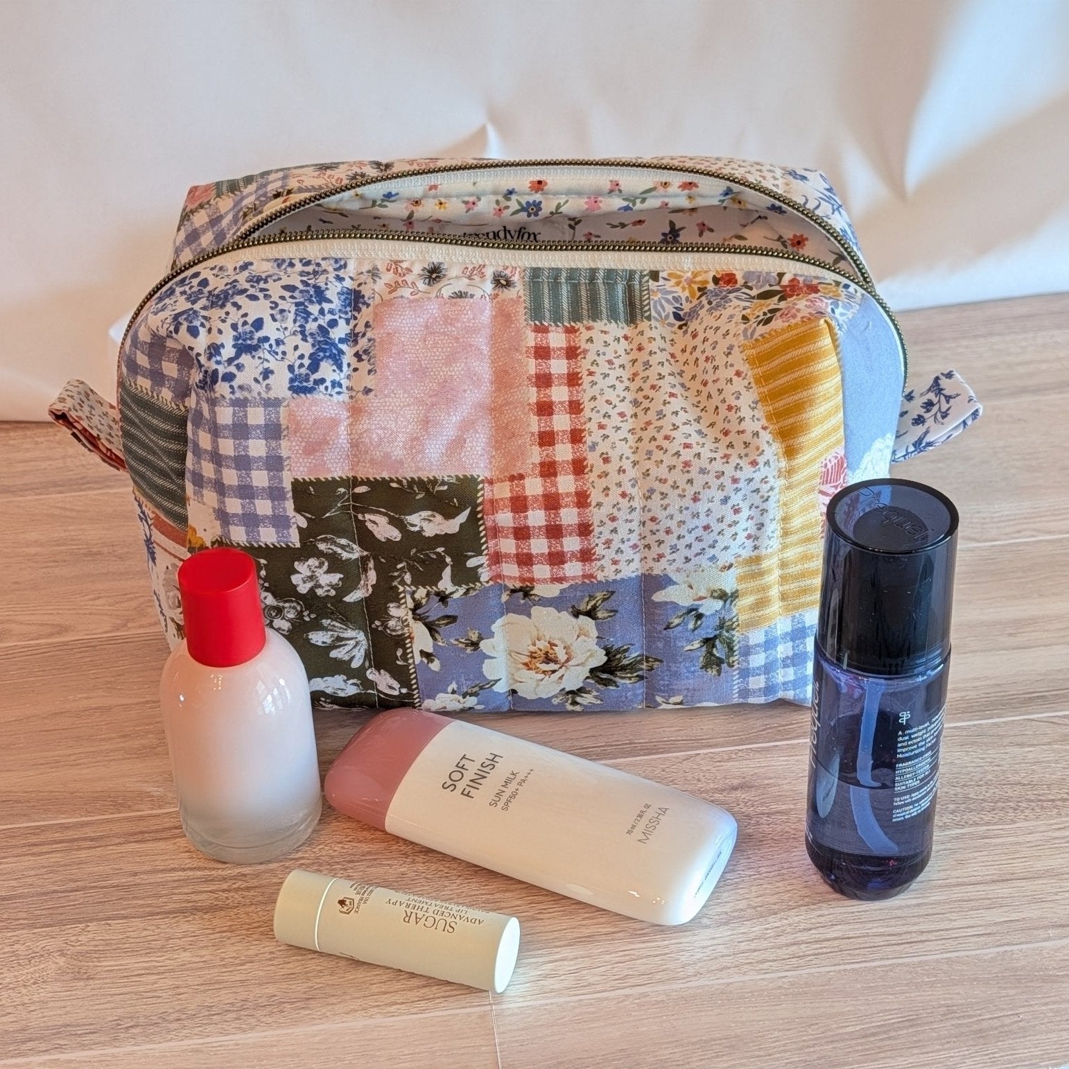 A patchwork fabric cosmetic pouch with a curved top and zip closure, displayed on a gray wood floor next to a potted green plant against a white paneled wall.
