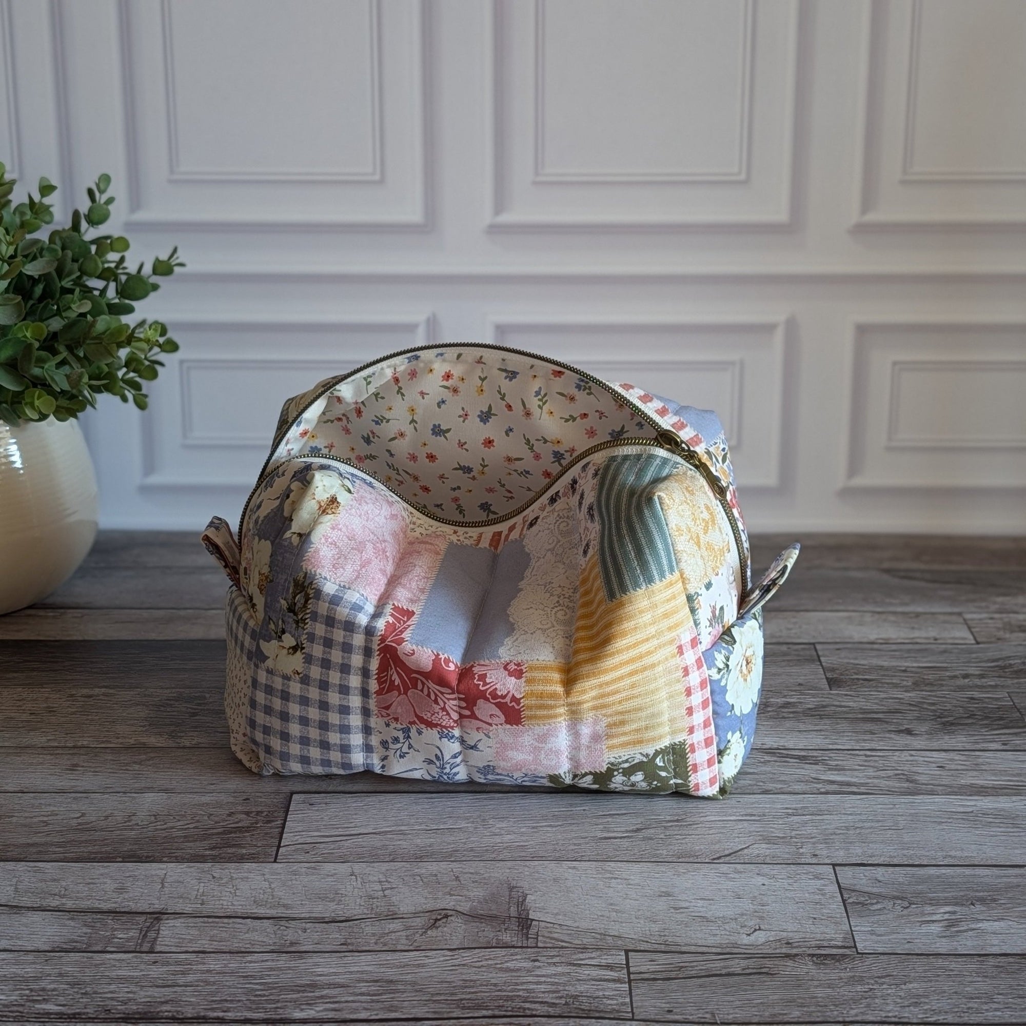 A patchwork fabric cosmetic pouch displayed on a gray wood floor next to a potted green plant against a white paneled wall.