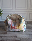 A patchwork fabric cosmetic pouch displayed on a gray wood floor next to a potted green plant against a white paneled wall.