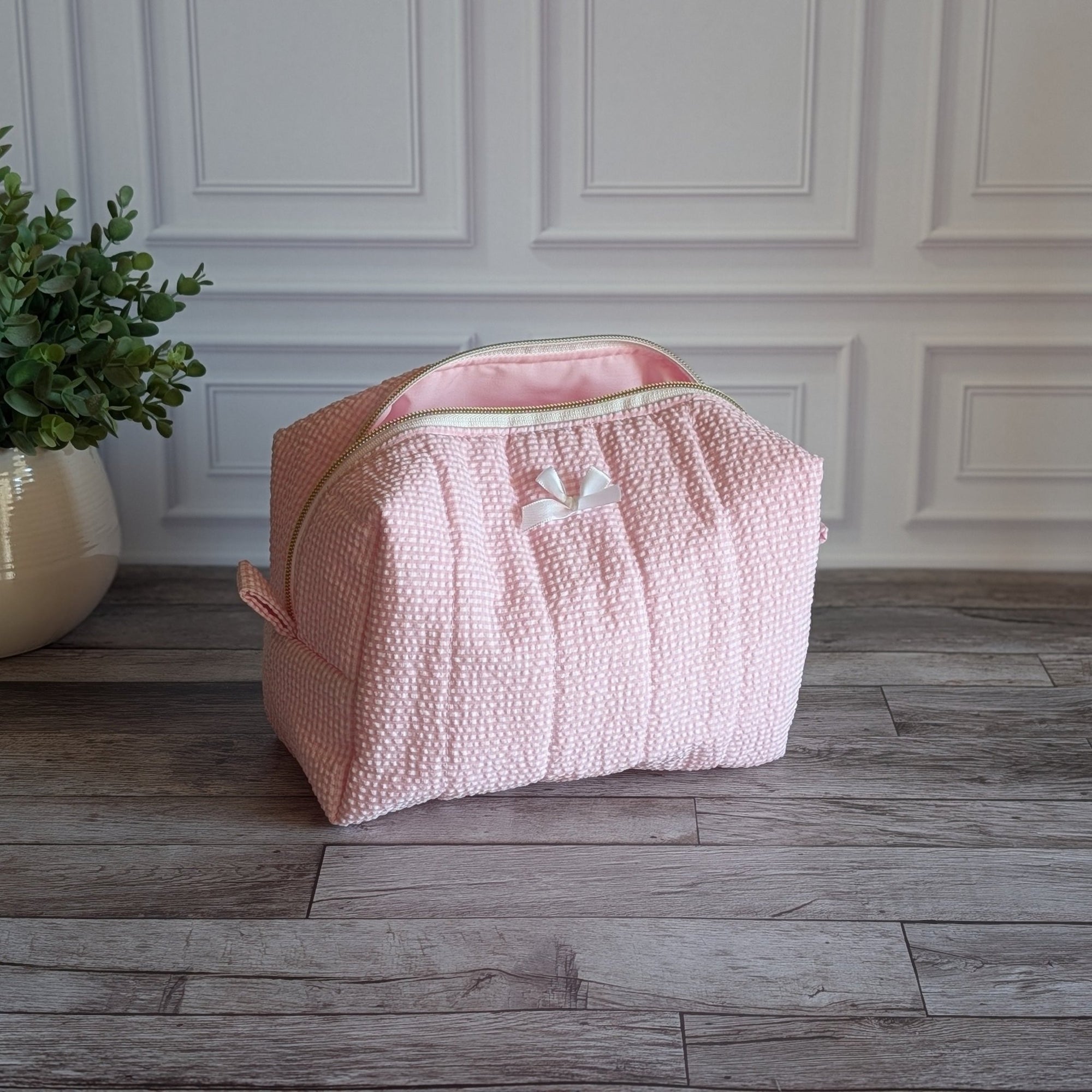 A light pink quilted cosmetic bag with a small white bow detail, sitting on a gray wood floor in front of a white paneled wall with a potted plant on the left.