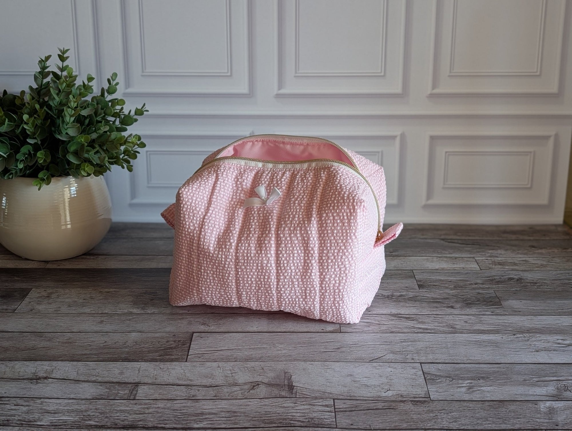 A light pink textured fabric storage bag or organizer with a zippered top, placed on a gray wood floor against a white paneled wall, next to a potted green plant.
