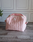 A light pink textured fabric storage bag or organizer with a zippered top, placed on a gray wood floor against a white paneled wall, next to a potted green plant.
