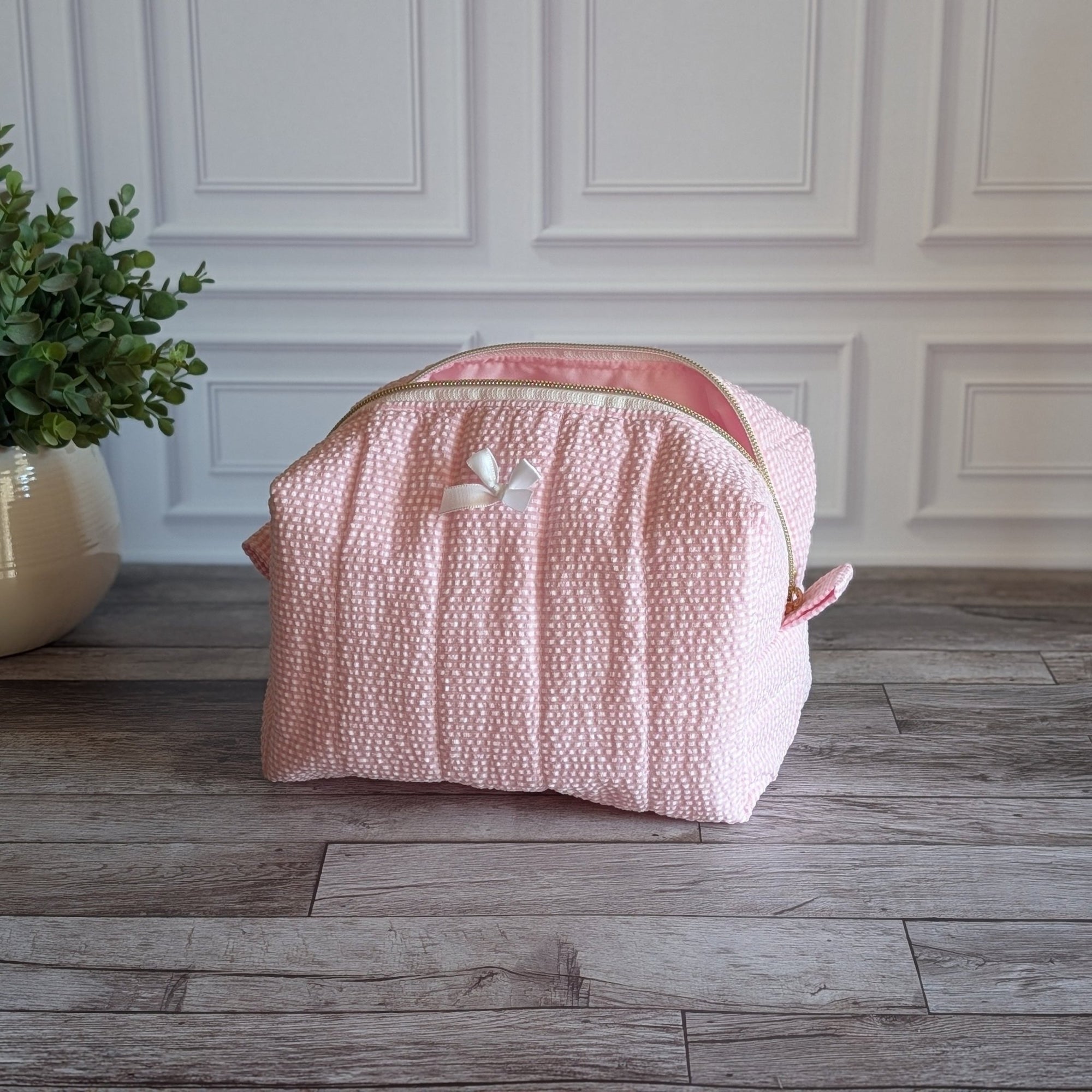 A light pink textured cosmetic bag with a white bow accent, sitting on a gray wood floor in front of a white paneled wall with a potted plant on the left.