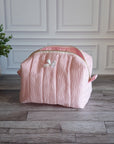 A light pink textured cosmetic bag with a white bow accent, sitting on a gray wood floor in front of a white paneled wall with a potted plant on the left.