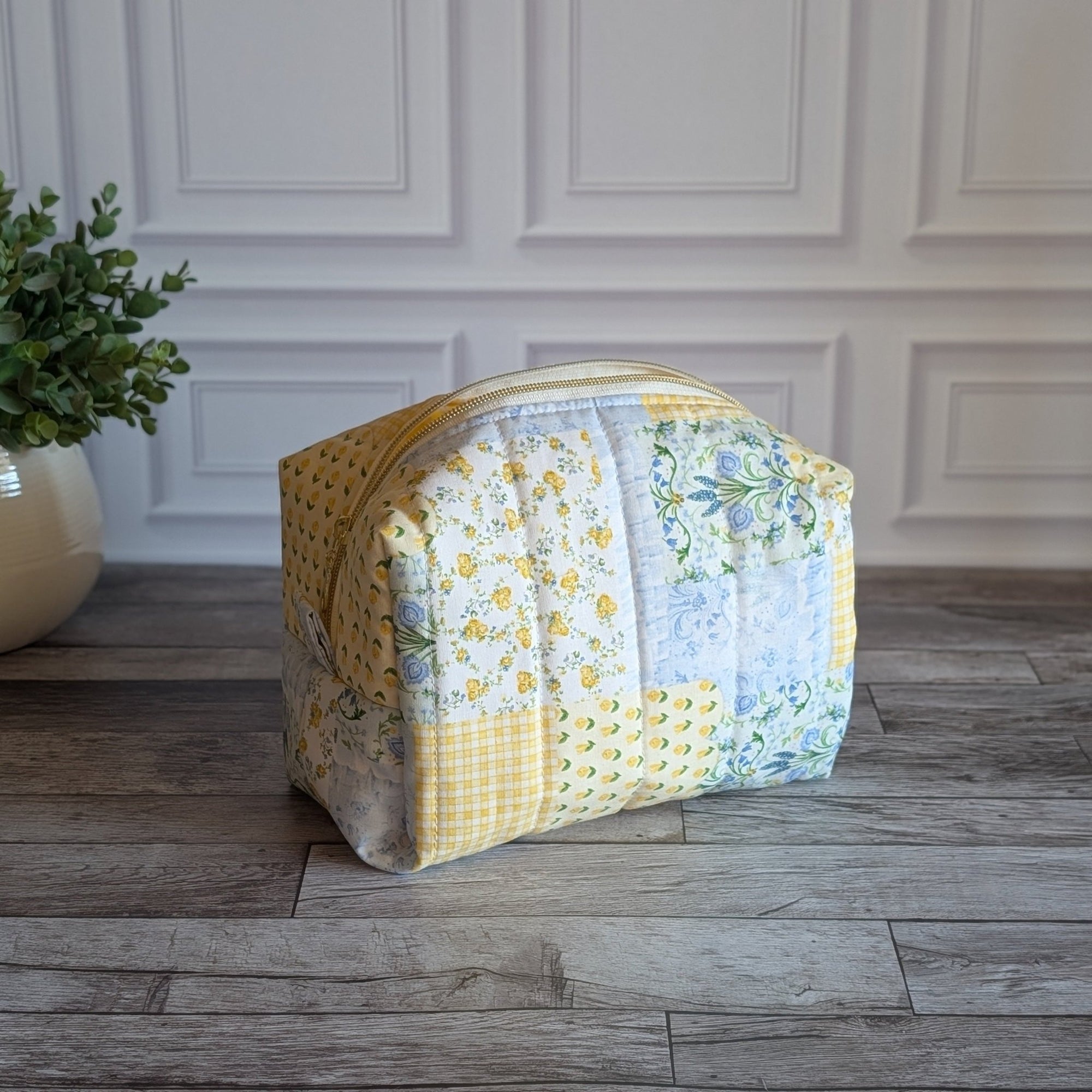A handmade patchwork cosmetic bag made from yellow, white, and floral fabrics, sitting on a gray wood floor in front of a white paneled wall with a potted plant on the left.