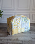 A handmade patchwork cosmetic bag made from yellow, white, and floral fabrics, sitting on a gray wood floor in front of a white paneled wall with a potted plant on the left.