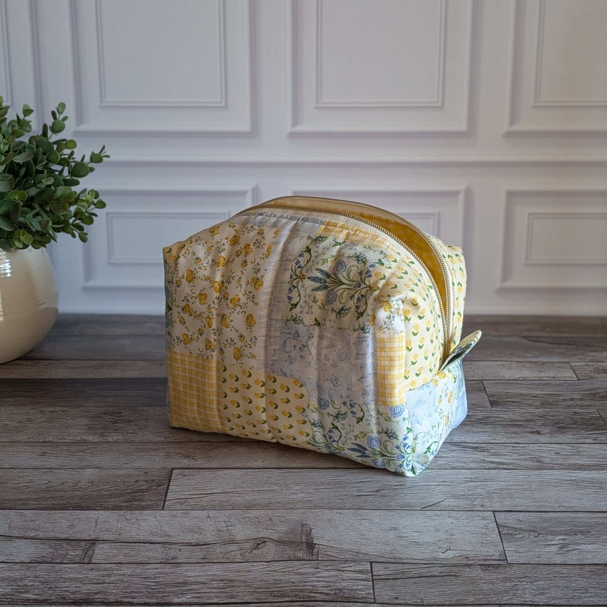 A quilted fabric zippered pouch in a yellow and floral patchwork pattern, sitting on a gray wood floor in front of a white paneled wall with a potted plant on the left.
