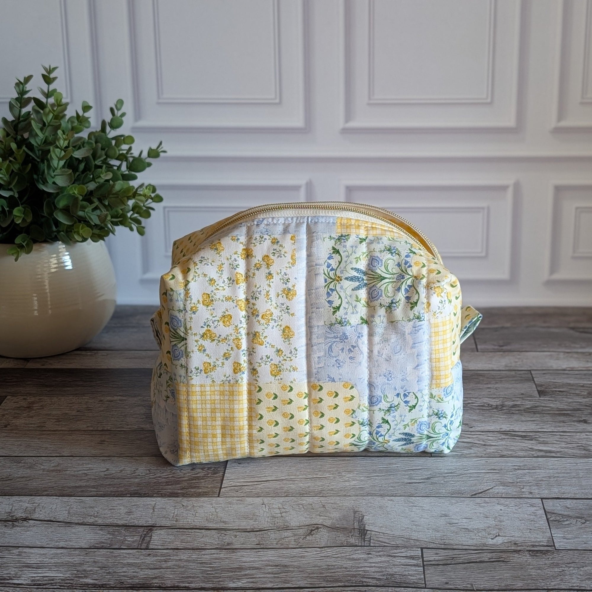 A quilted patchwork cosmetic bag in yellow, white, and floral prints, sitting on a gray wood floor next to a potted green plant against a white paneled wall.