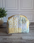A quilted patchwork cosmetic bag in yellow, white, and floral prints, sitting on a gray wood floor next to a potted green plant against a white paneled wall.