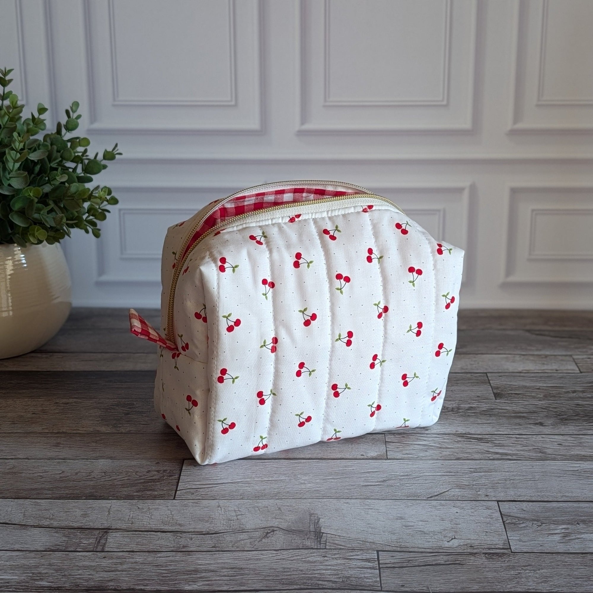 A quilted white cosmetic bag with a red and white cherry print and a red gingham zipper, sitting on a gray wood floor next to a potted green plant.