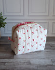 A quilted white cosmetic bag with a red and white cherry print and a red gingham zipper, sitting on a gray wood floor next to a potted green plant.
