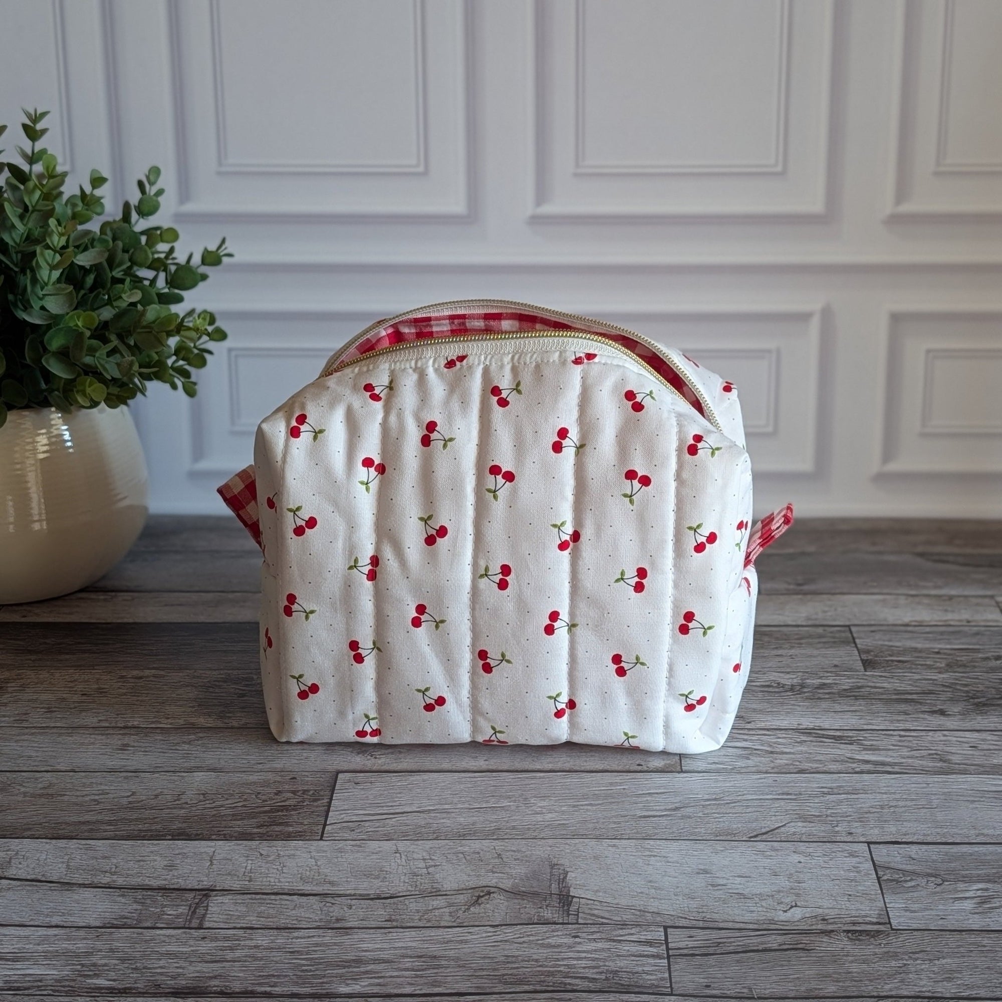 A quilted white cosmetic bag with a red cherry print and red gingham trim, sitting on a gray wood floor next to a potted green plant.