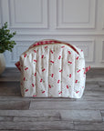 A quilted white cosmetic bag with a red cherry print and red gingham trim, sitting on a gray wood floor next to a potted green plant.