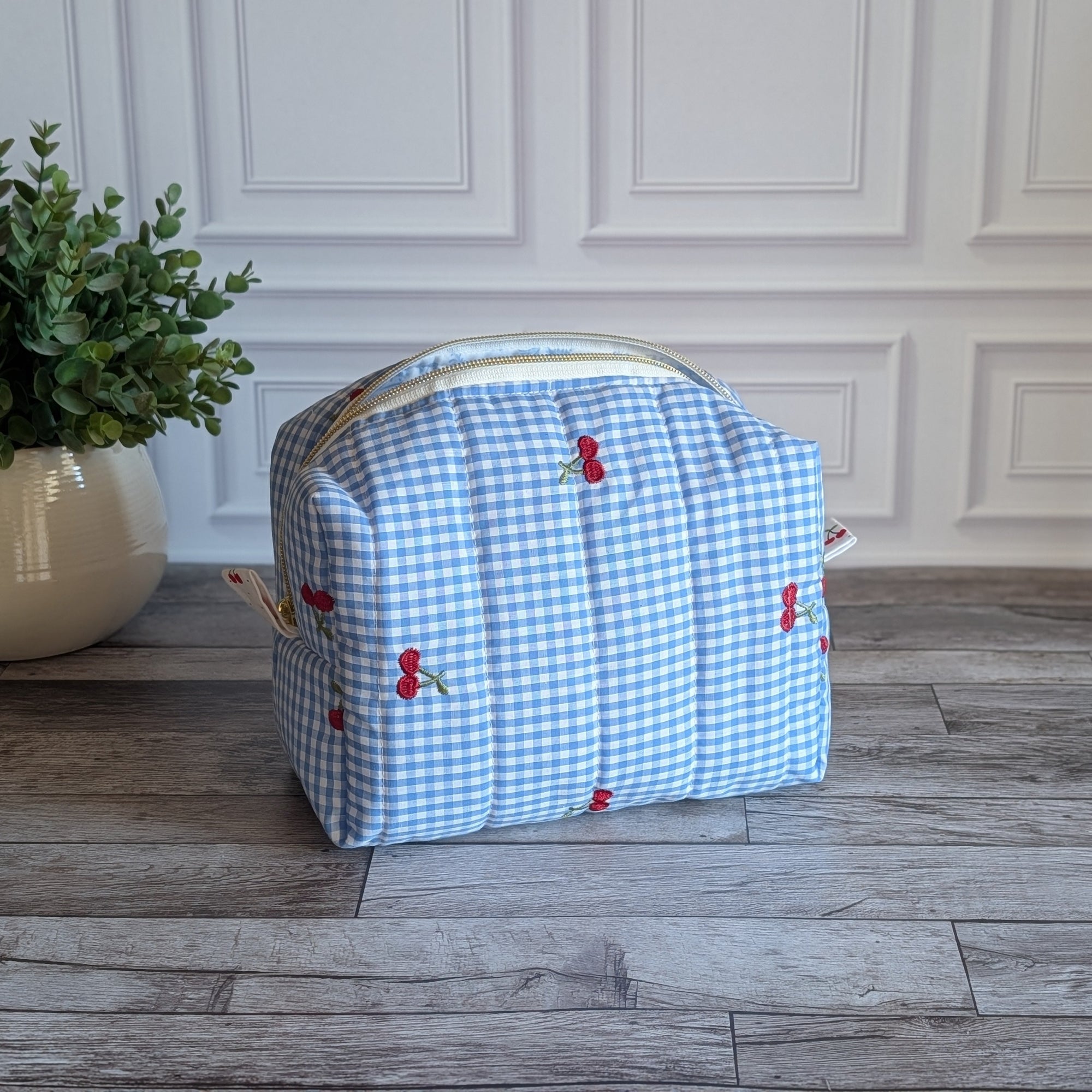 A light blue and white gingham toiletry bag with small red cherub motifs, resting on a gray wood plank floor in front of a white paneled wall next to a potted green plant.
