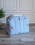 A light blue and white gingham toiletry bag with small red cherub motifs, resting on a gray wood plank floor in front of a white paneled wall next to a potted green plant.