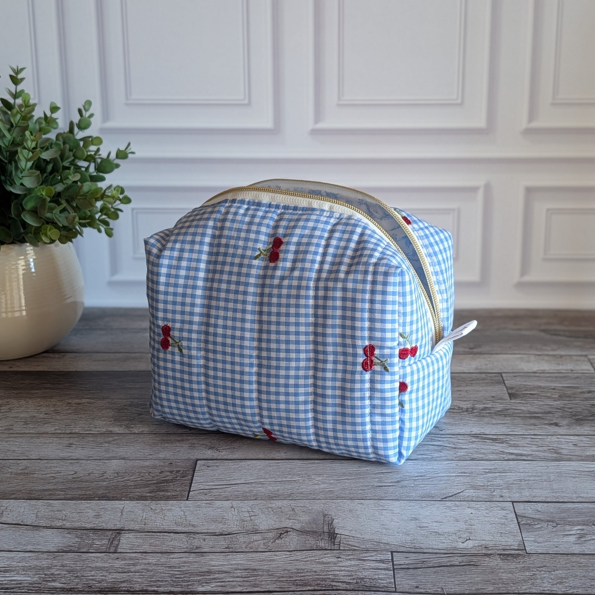A light blue and white gingham toiletry bag with small red embroidered cherries, sitting on a gray wood floor in front of a white paneled wall.