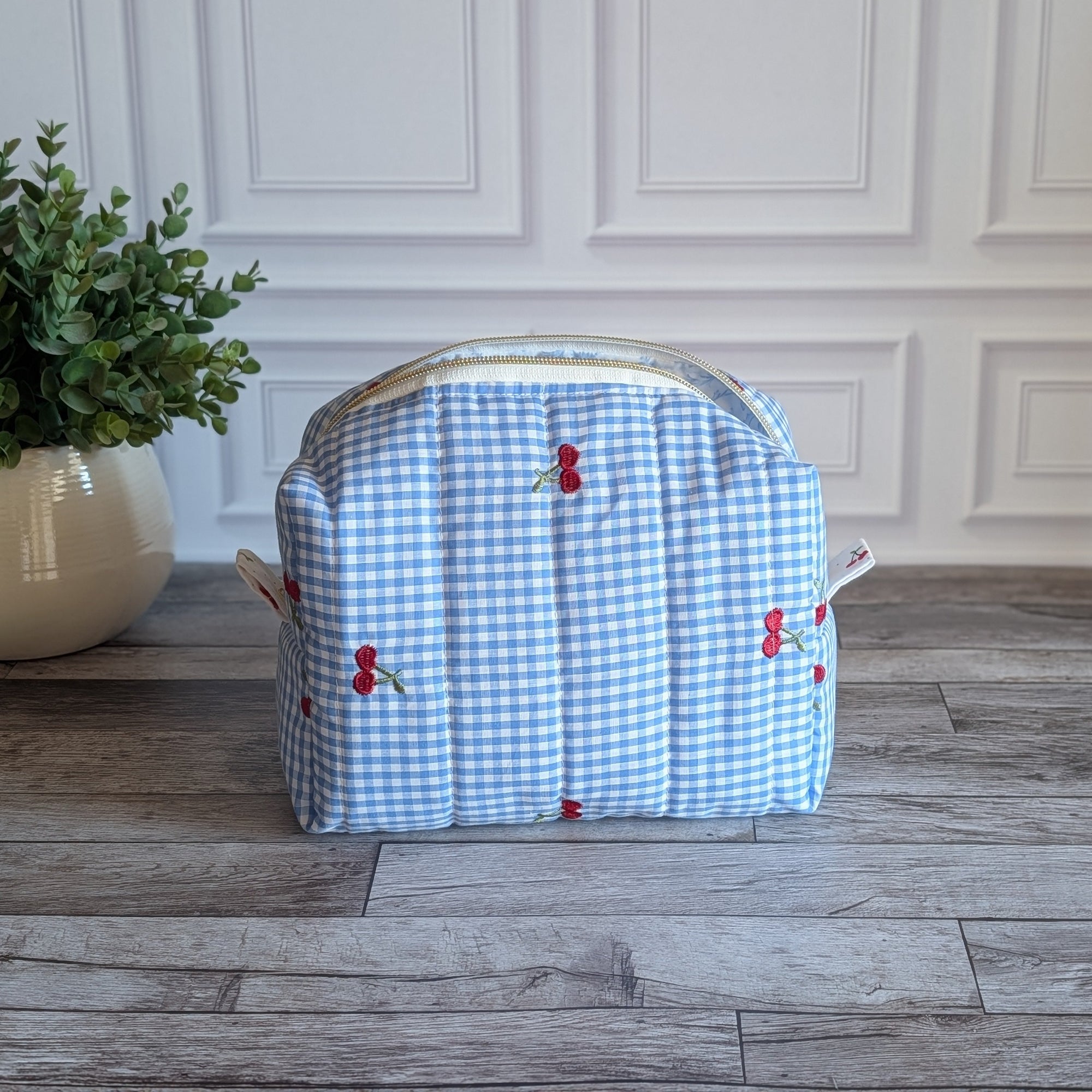 A light blue and white gingham toiletry bag with red cherries and green leaves embroidered on the front, sitting on a gray wood floor in front of a white paneled wall.
