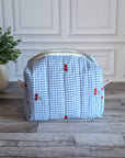 A light blue and white gingham toiletry bag with red cherries and green leaves embroidered on the front, sitting on a gray wood floor in front of a white paneled wall.
