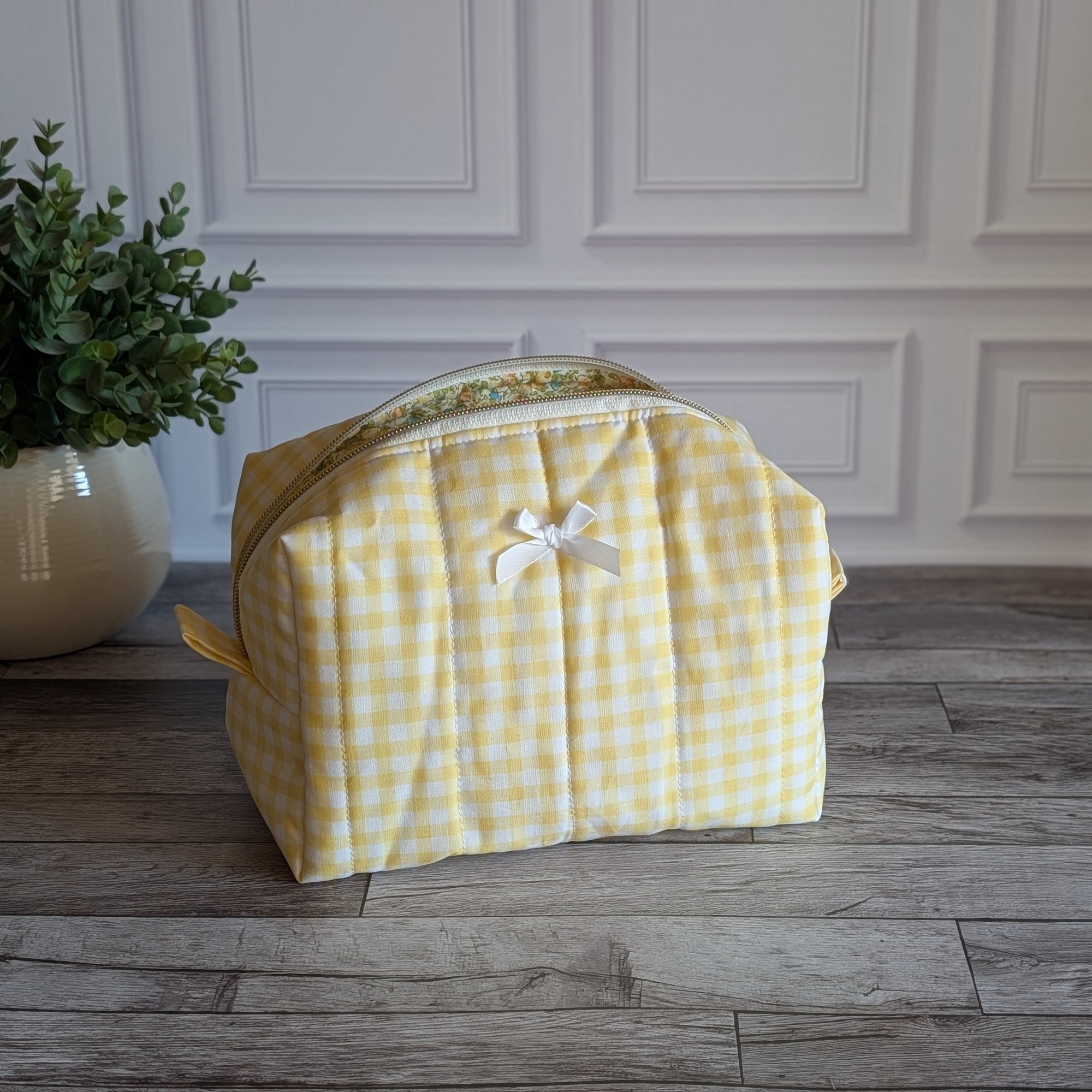 A yellow and white gingham quilted toiletry bag with a small white bow on the front, set on a gray wood floor against a white paneled wall with a potted plant on the left.